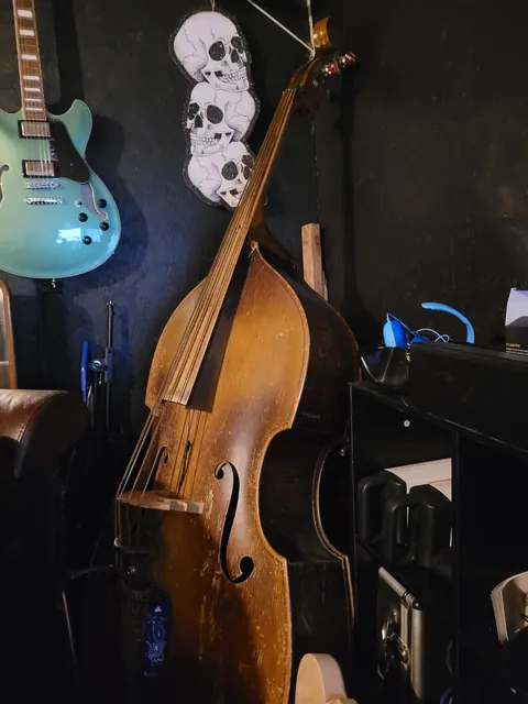 A well-worn upright bass leaning in the corner of the shop, with a skull decoration and a seafoam green hollow-body guitar hanging on the black wall.