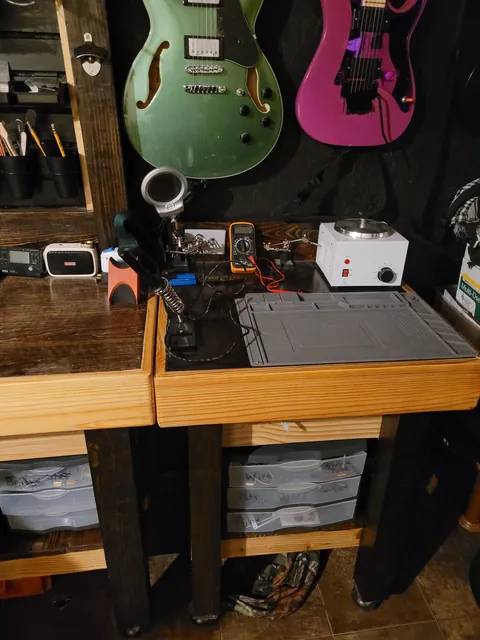 A workbench with a soldering station and tools, with a green hollow-body guitar and a magenta guitar hanging behind it.