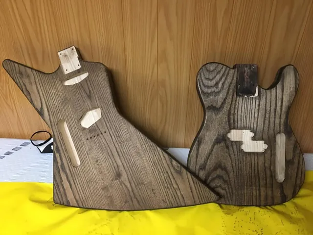 Two unfinished ash guitar bodies, an explorer shape and a Tele shape, with routed pickup cavities.