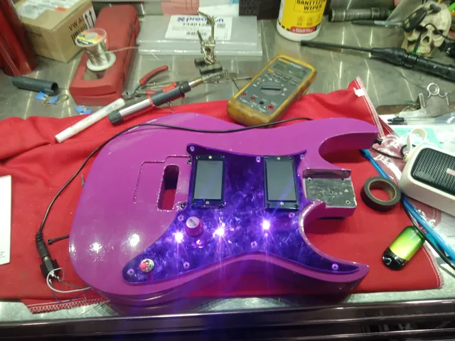 A purple guitar body with a mirrored purple pickguard and two humbuckers, being wired on a red shop towel, with a multimeter and soldering iron nearby.